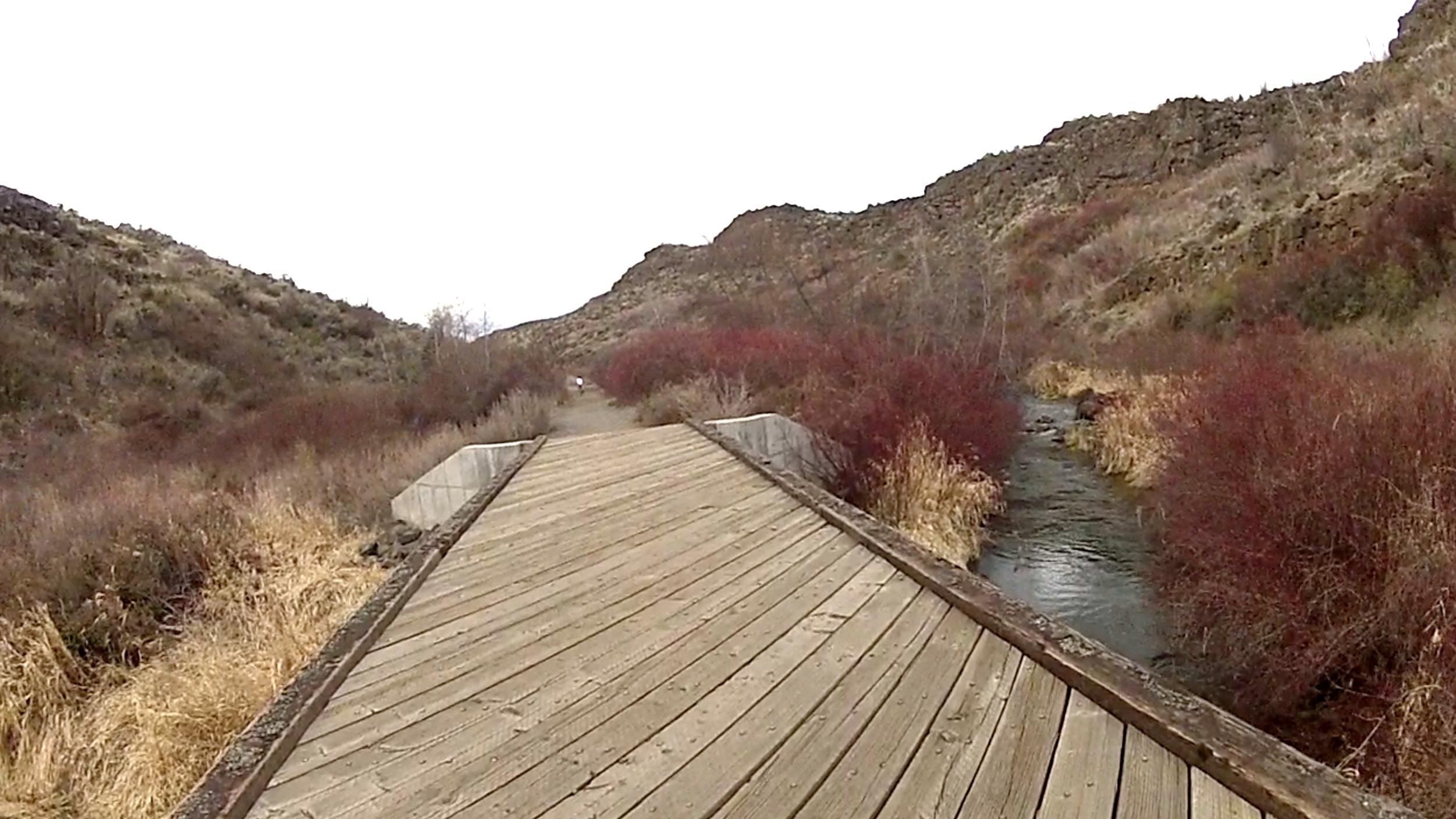 A wooden bridge extending over a flowing creek, surrounded by hilly terrain and patches of reddish-brown brush. The sky is overcast, creating a serene atmosphere along the pathway leading into the distance. Cowiche Canyon mountain bike trail.