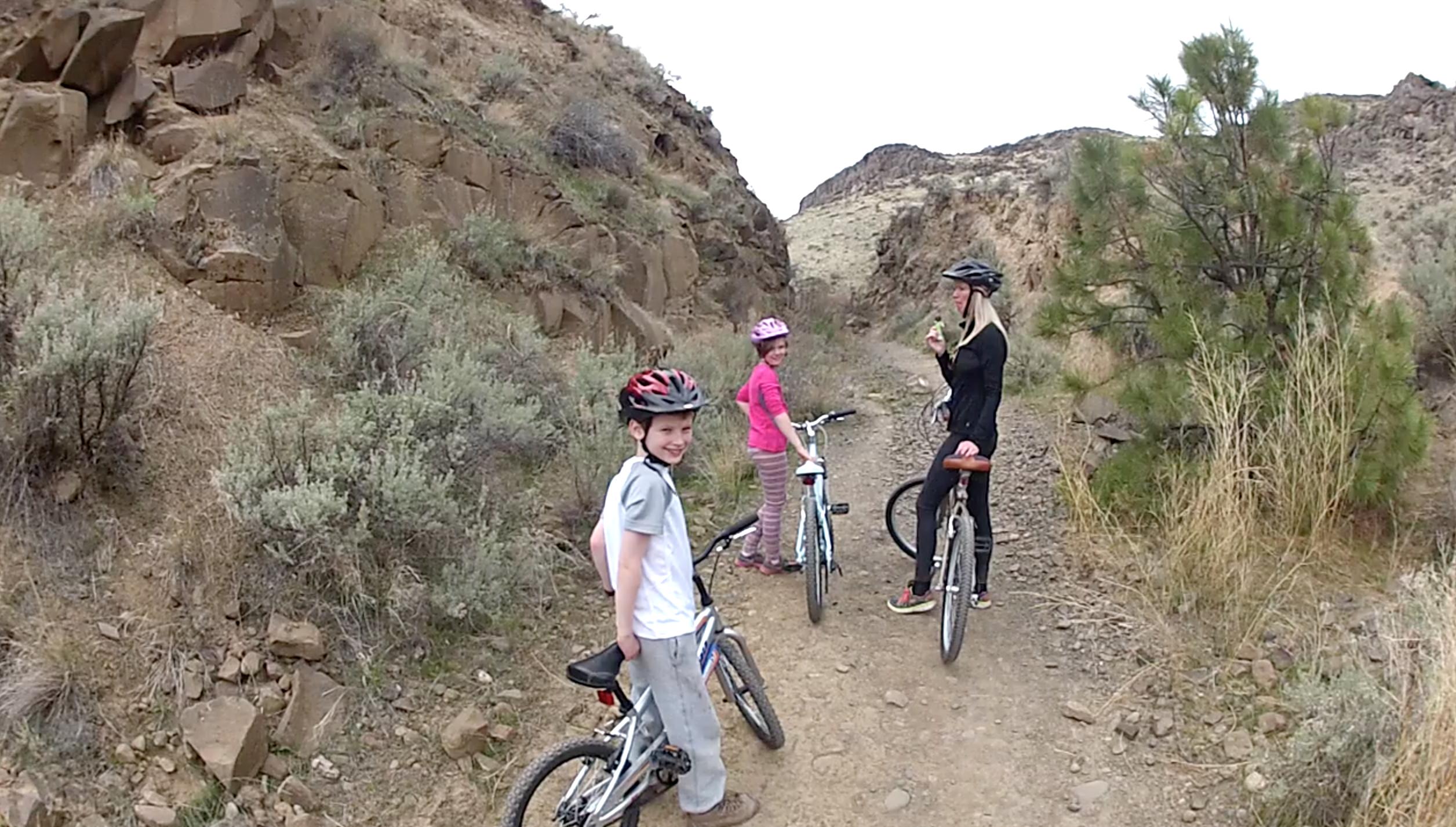Three children and an adult are enjoying a break while biking on a rugged trail. The background features rocky terrain and sparse vegetation. The boy in the foreground is smiling at the camera, while two girls, one in a pink shirt and one in a striped outfit, stand beside their bikes. The adult, dressed in black, is eating an ice cream and looks back, contributing to the cheerful atmosphere of their outdoor adventure. Cowiche Canyon mountain bike trail.