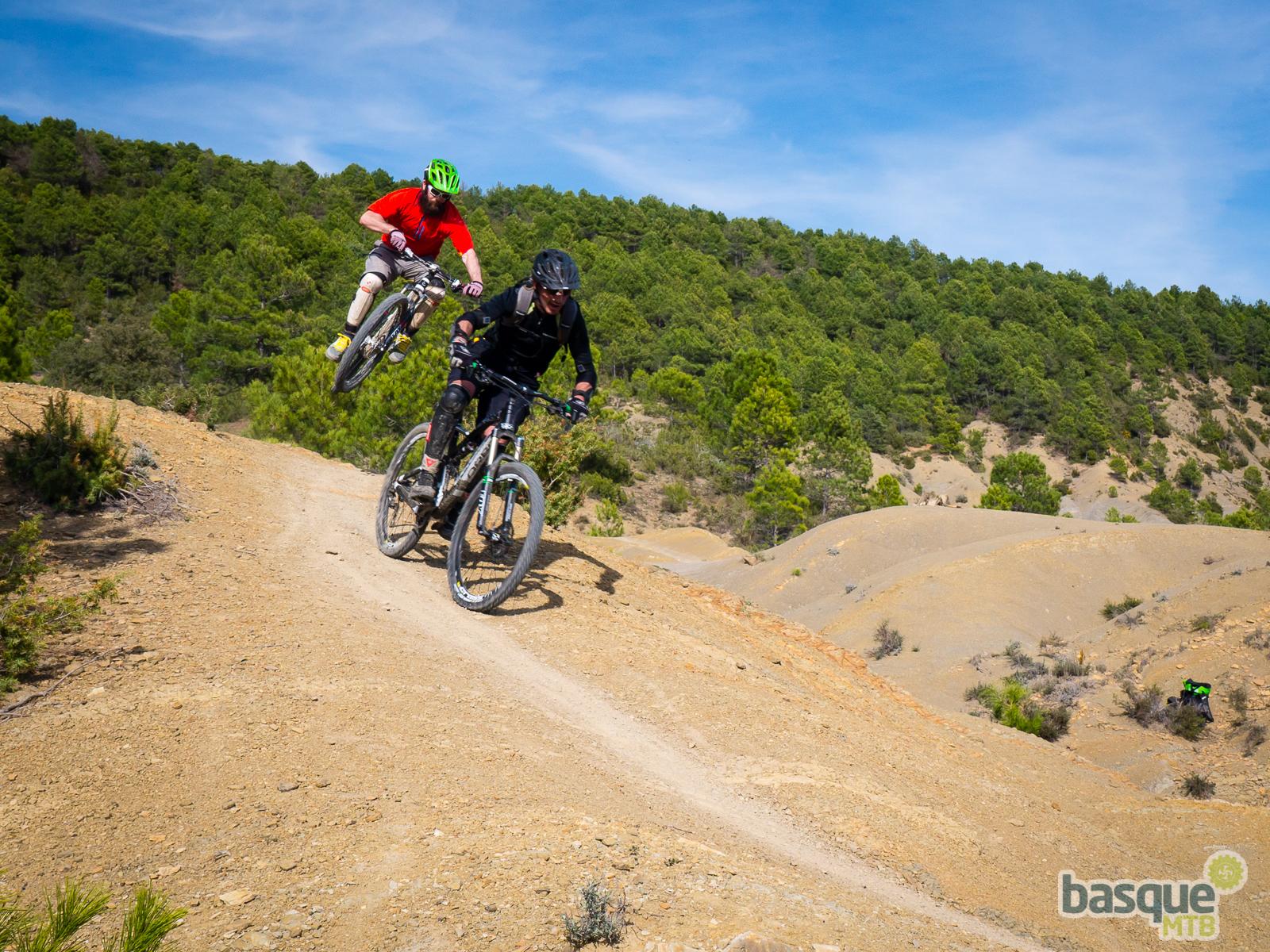 Orbea Occam 29 H10: Two mountain bikers navigate a dusty trail in a lush green landscape. One rider, wearing a red shirt and a green helmet, is airborne, performing a jump, while the other rider in black gear is pedaling along the ground. The scene captures the thrill of mountain biking against a backdrop of soft hills and trees under a blue sky.