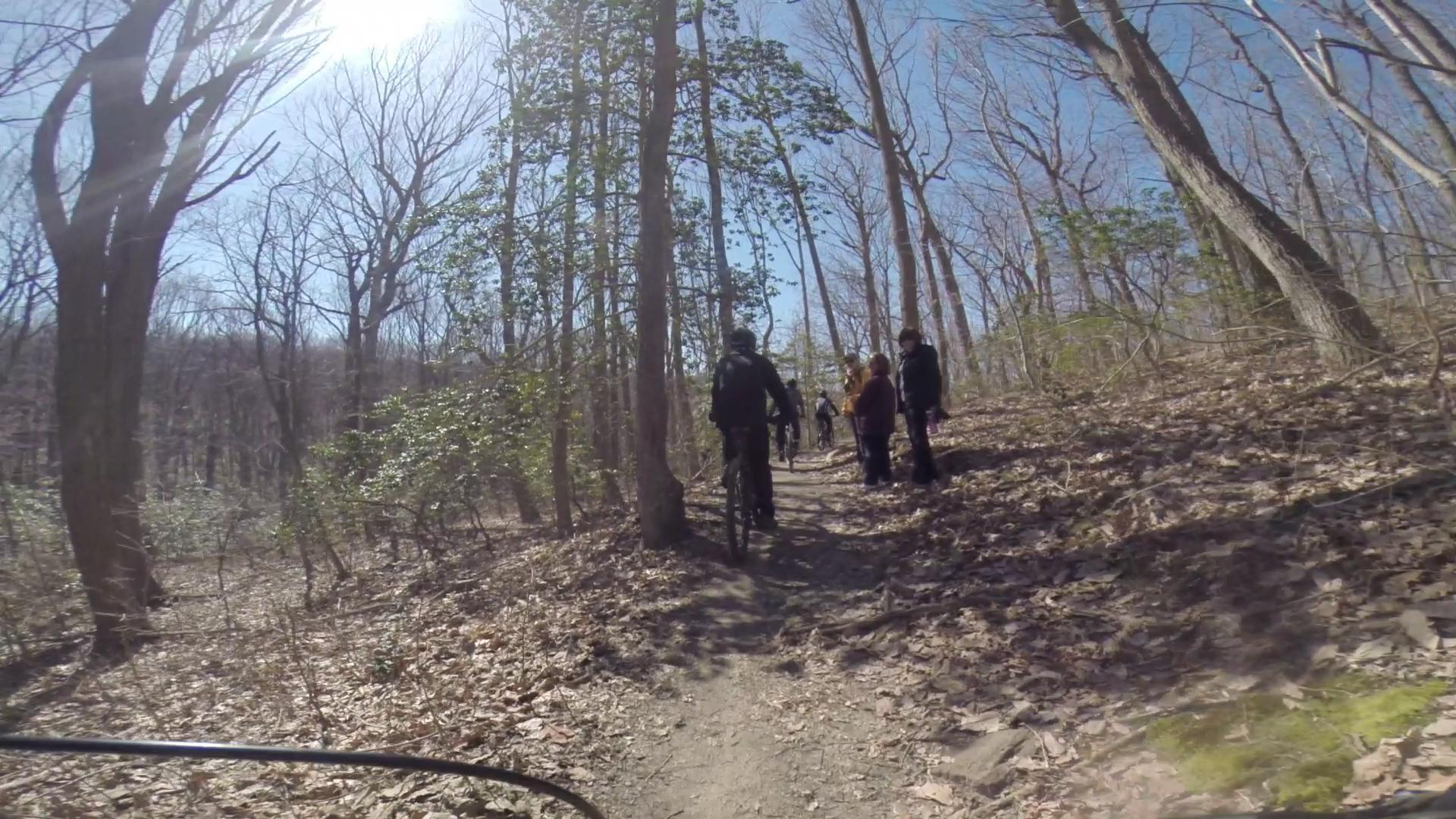 A group of people walking along a dirt trail in a wooded area, with bare trees and scattered leaves on the ground. Sunlight filters through the branches, creating a bright atmosphere on a clear day. Hartshorne Woods Park mountain bike trail.