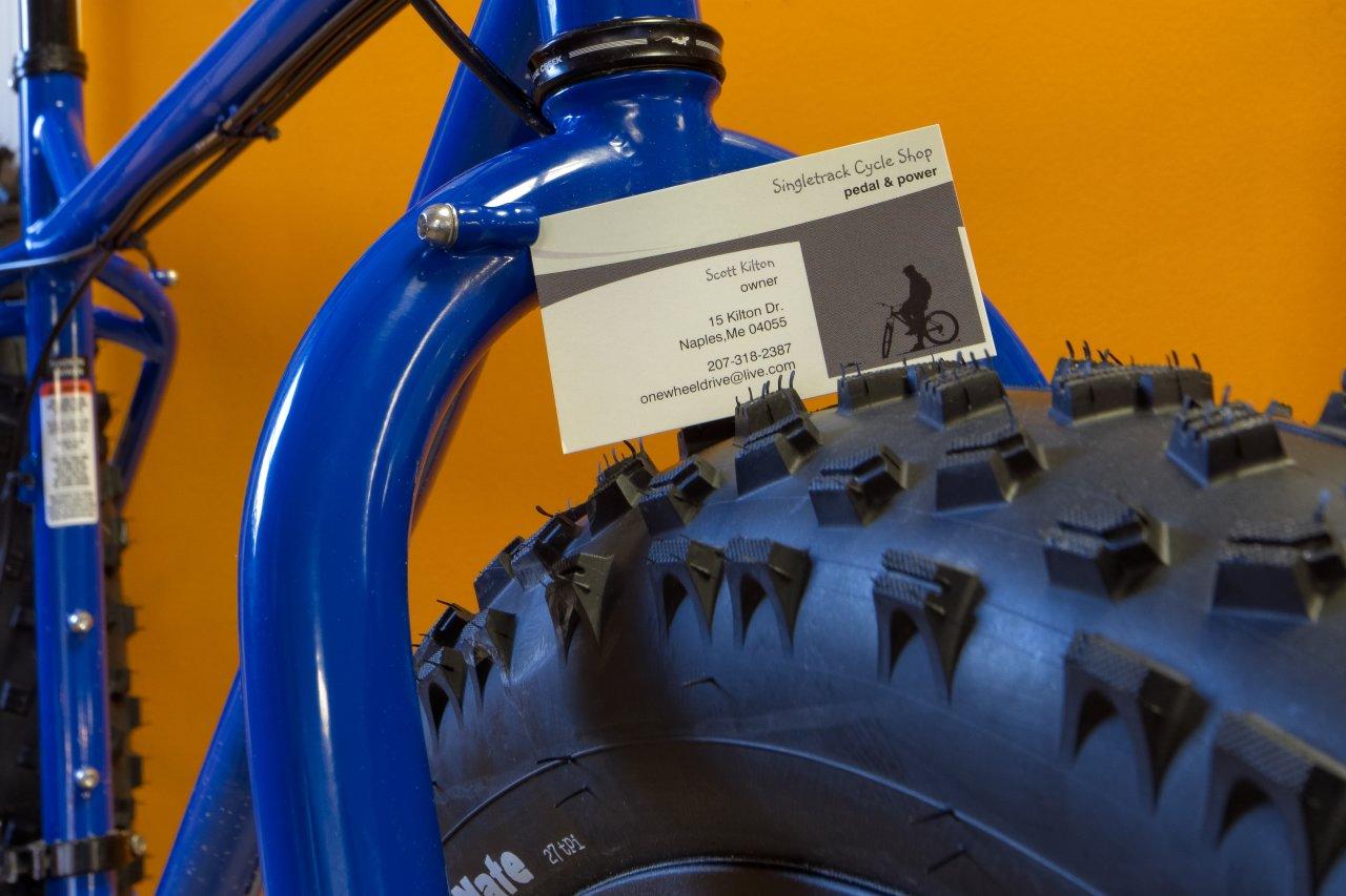 A close-up image of a blue bicycle frame with a business card for "Singletrack Cycle Shop" placed against a fat tire. The background is a bright orange. The card includes the owner's name, contact information, and address.