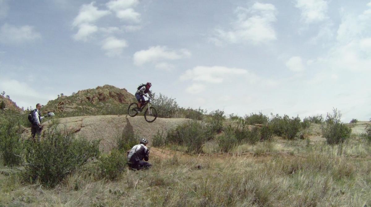 Santa Cruz Nomad Carbon: A mountain biker performing a jump over a rocky terrain, while two onlookers watch from the side in a grassy, natural environment under a partly cloudy sky.