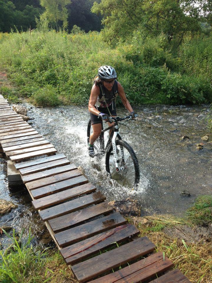 A cyclist rides a mountain bike across a wooden bridge over a stream, splashing water as they navigate the terrain. Lush greenery surrounds the area, creating a scenic outdoor setting. The sun is shining, suggesting a bright and clear day.