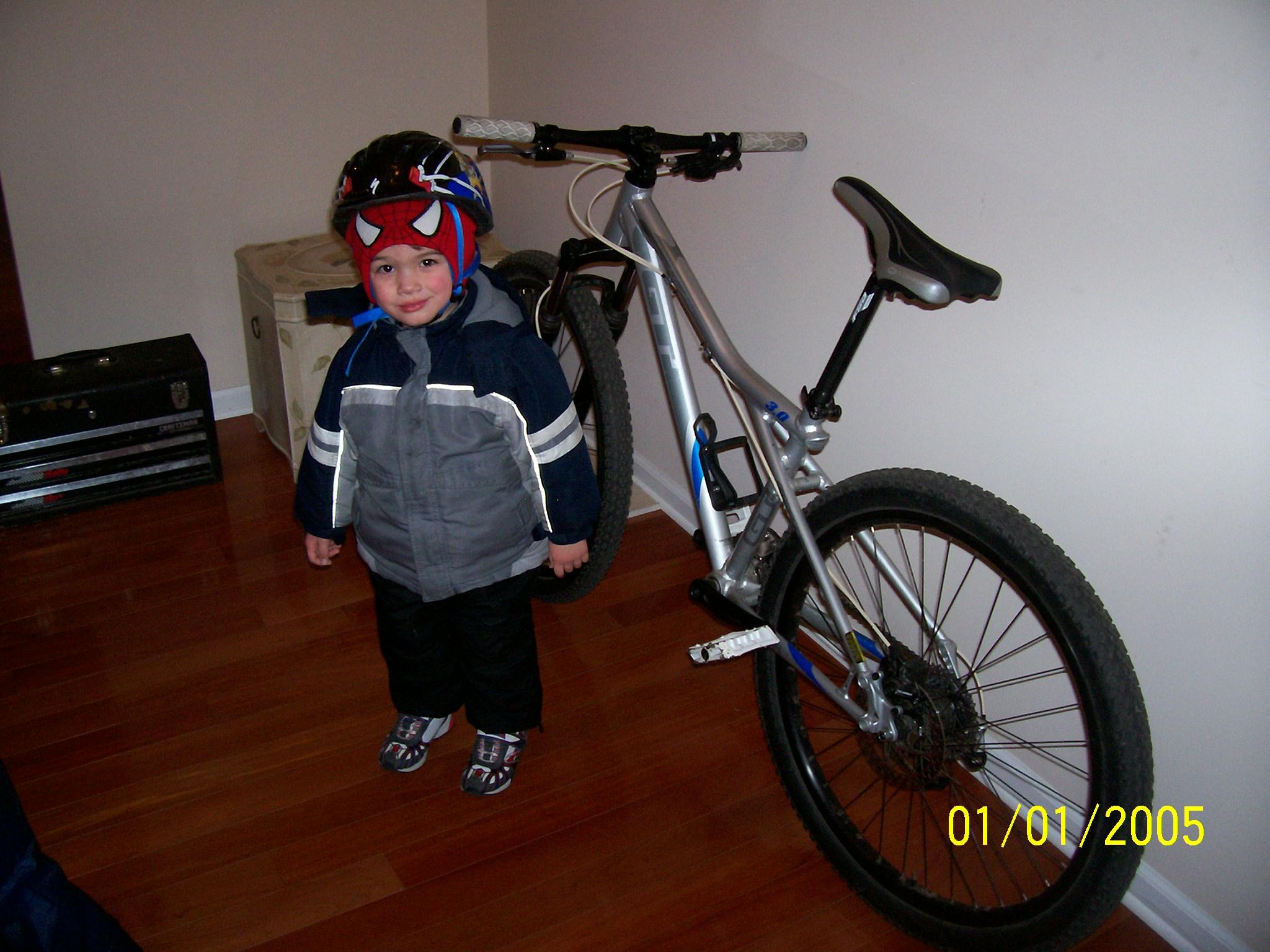 GT Avalanche: A young child wearing a Spider-Man-themed helmet and a warm jacket stands next to a mountain bike in a home setting. The child is smiling and positioned on a wooden floor, with a storage box visible in the background. The bike has a silver frame and is propped against the wall.