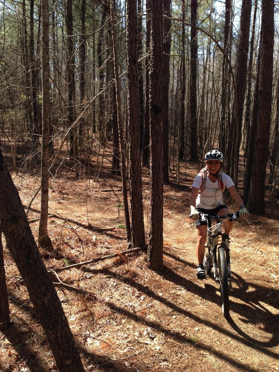 A person riding a mountain bike along a narrow dirt trail in a forest, surrounded by tall trees. Sunlight filters through the branches, illuminating the dirt path covered with pine needles. The rider is wearing a helmet and a light-colored shirt, with a backpack visible. Harbins Park mountain bike trail.