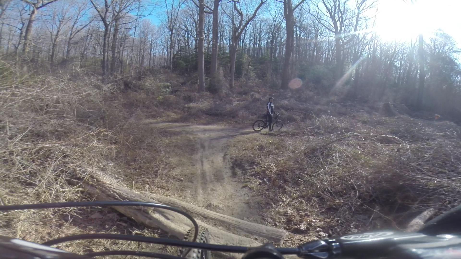 A mountain biker stands next to their bike on a dirt trail surrounded by a wooded area with bare trees and brush. Sunlight peeks through the branches, illuminating the scene. The trail curves ahead and there is a log across the path. Hartshorne Woods Park mountain bike trail.