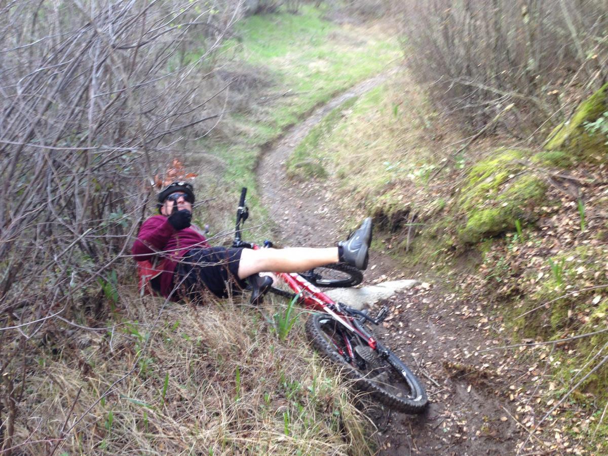 A person lying on the ground next to a mountain bike, surrounded by bushes and grass, giving a thumbs-up gesture. The background shows a winding dirt trail through a natural setting. Granite Bay Trail mountain bike trail.