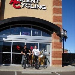 Two men standing in front of a bicycle shop called "Ascent Cycling." They are each holding a bicycle, and the store has large glass doors with an "OPEN" sign displayed. The weather looks clear and sunny.