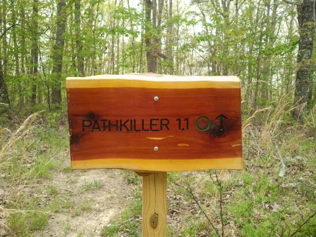 Wooden trail sign reading "PATHKILLER 1.1" with an arrow pointing up, surrounded by a wooded area and grassy terrain. Five Points mountain bike trail.