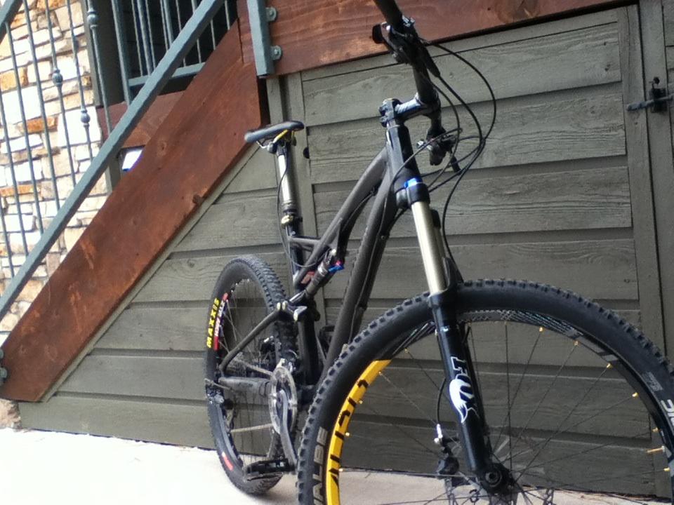 Specialized Stumpjumper FSR Expert Evo: A close-up view of a mountain bike positioned against a wooden wall. The bike features black and silver components, including a front suspension fork and knobby tires with yellow accents. The background includes wooden steps and a stone wall, indicating an outdoor setting.