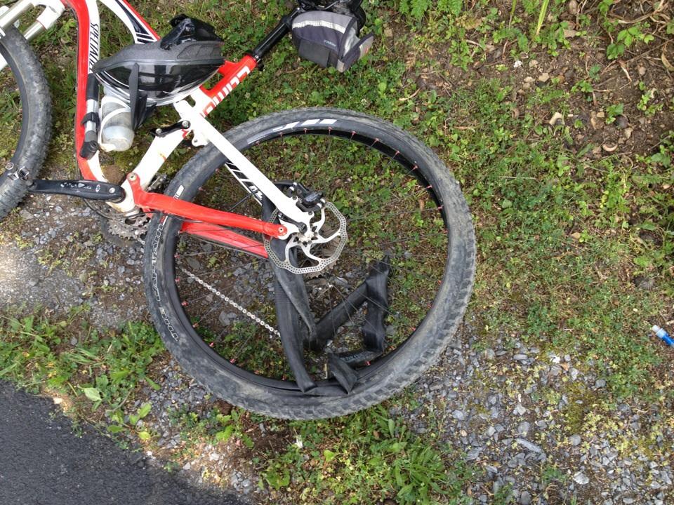 Specialized Camber: A close-up view of a mountain bike laying on grass and gravel, showcasing the rear wheel and drivetrain components. The bike features a red and white frame, a helmet resting on the top tube, and a water bottle attached to the frame.