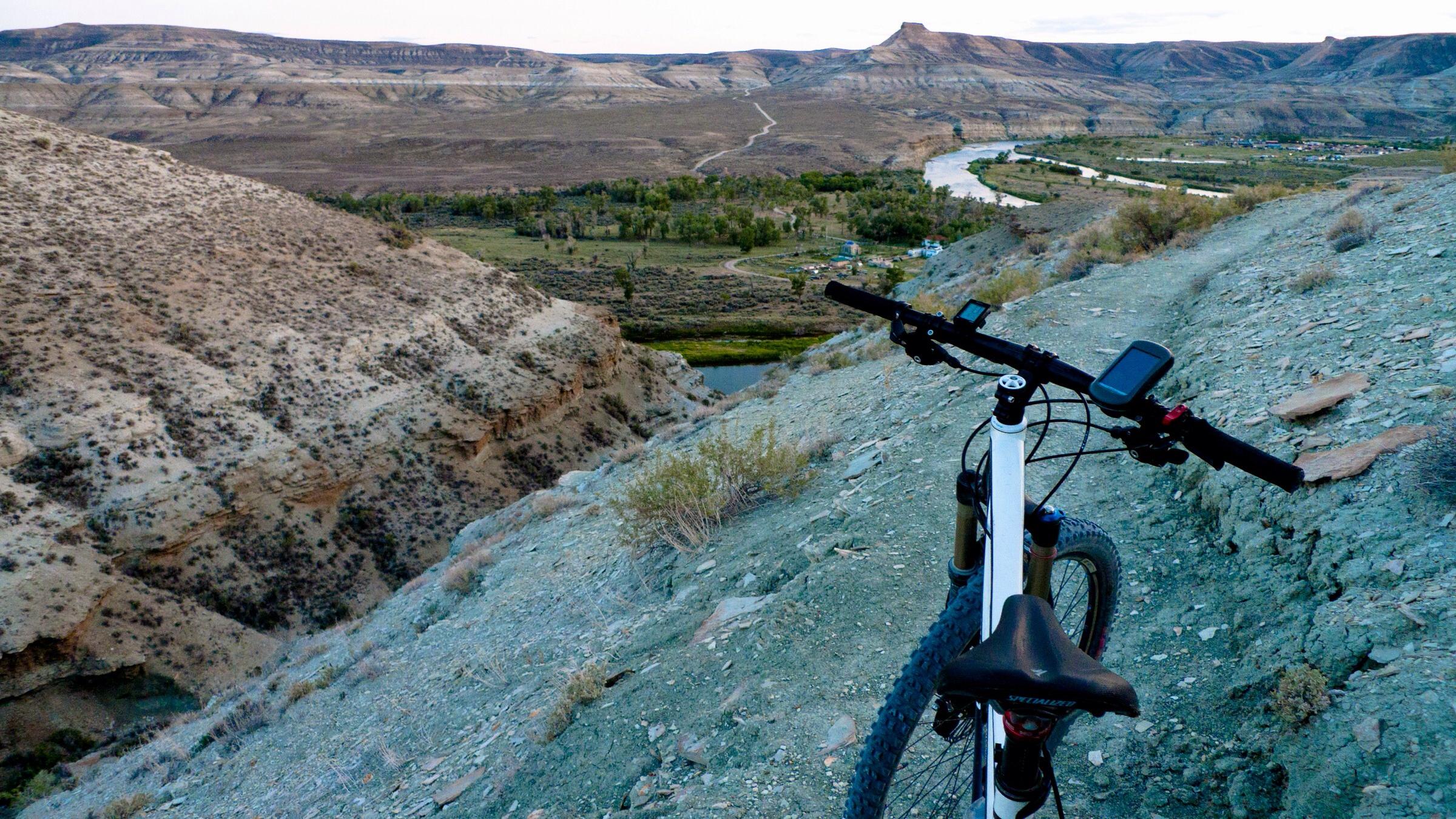 Specialized Stumpjumper FSR Comp 29: A mountain bike rests on a rugged hillside overlooking a winding river and valley below, framed by distant hills and rolling terrain. The landscape features dry earth with sparse vegetation, capturing a serene outdoor scene ideal for biking and exploration.