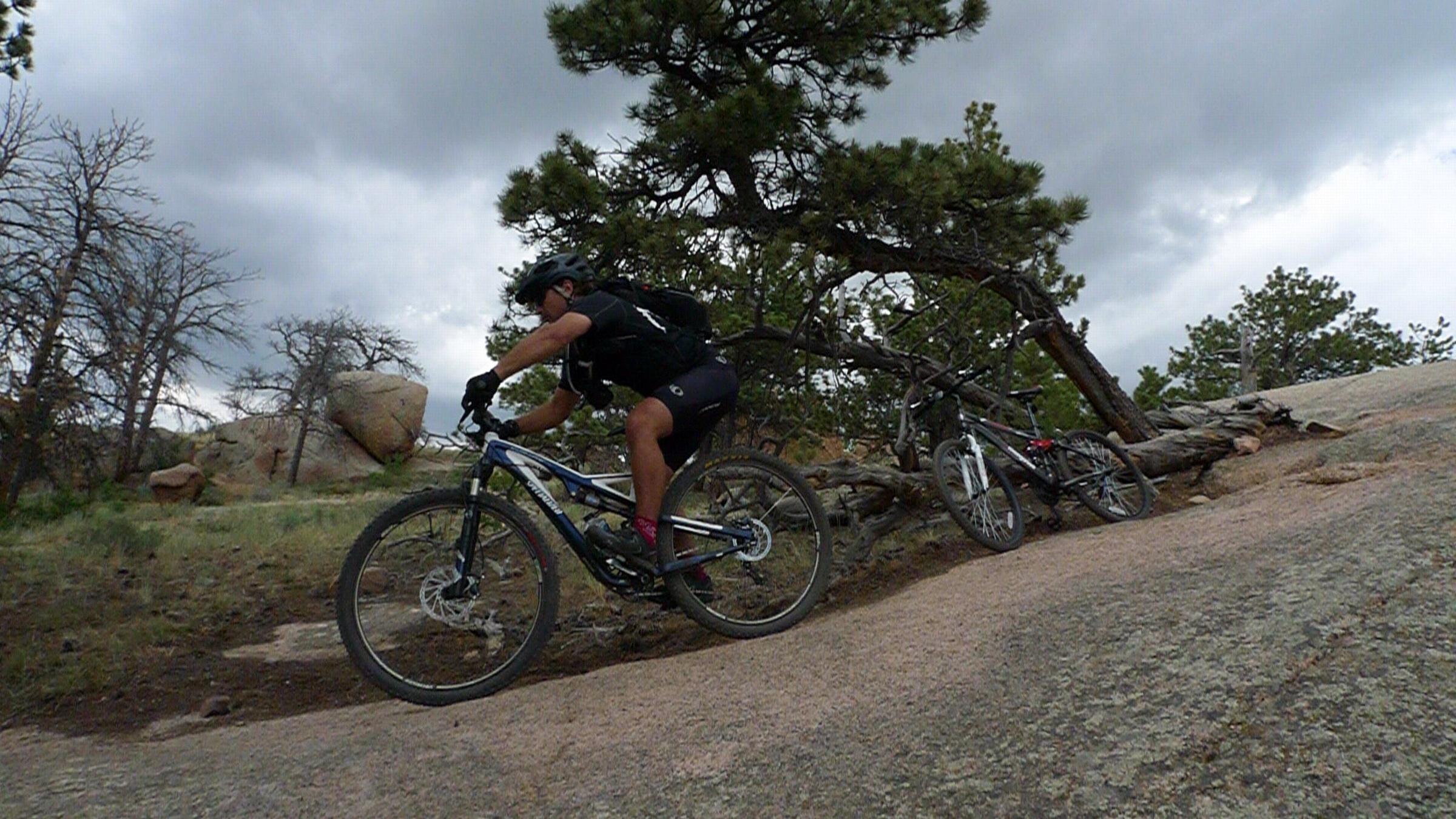 Specialized Stumpjumper FSR Comp 29: A mountain biker navigates a rocky trail surrounded by trees and boulders under a cloudy sky. Two additional bicycles are resting nearby on the trail.