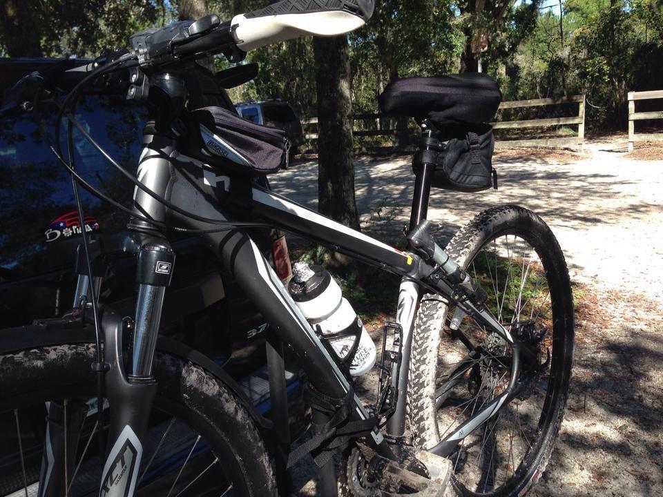 Specialized Stumpjumper FSR Comp 29: Mountain bike parked beside a vehicle, featuring a water bottle holder and a small bag on the seat, set against a backdrop of trees and a dirt trail.