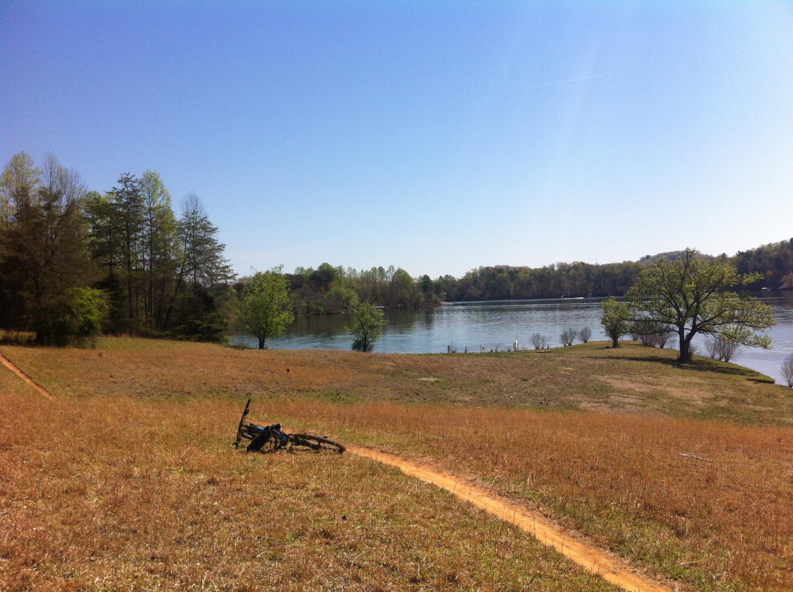 A peaceful outdoor scene featuring a grassy field sloping down to a calm lake. In the foreground, a bicycle lies on its side on a dirt path. The background includes lush trees and a clear blue sky, creating a tranquil atmosphere. Warrior Creek mountain bike trail.