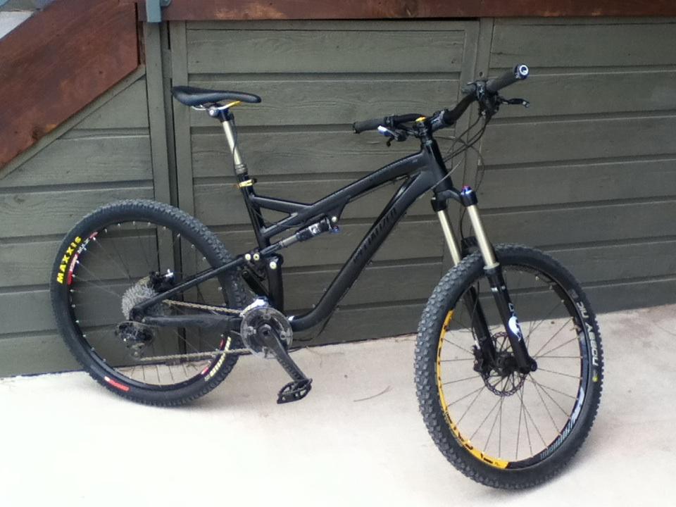 Specialized Stumpjumper FSR Expert Evo: A black mountain bike leaning against a wooden structure, featuring large, knobby tires and front suspension. The bike has a sleek design with prominent components, including a gear mechanism and handlebars, set against a muted green background.