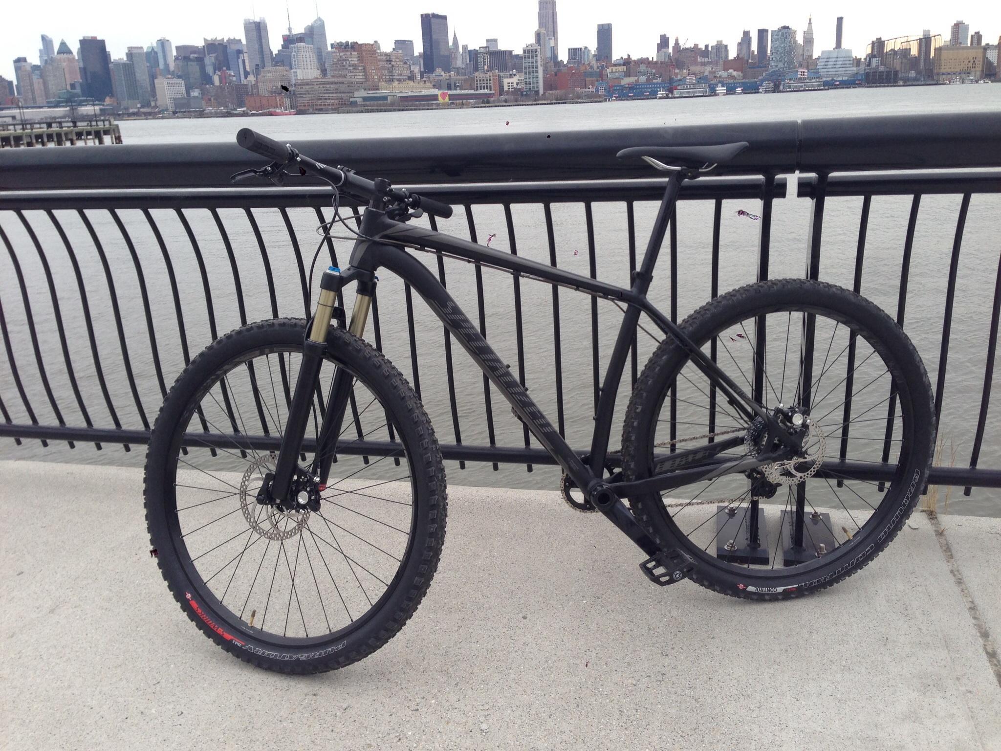 Specialized STUMPJUMPER EVO HT: A black mountain bike parked on a waterfront pathway, with a city skyline in the background. The bike features thick tires and a front suspension fork, positioned near a railing overlooking the water.