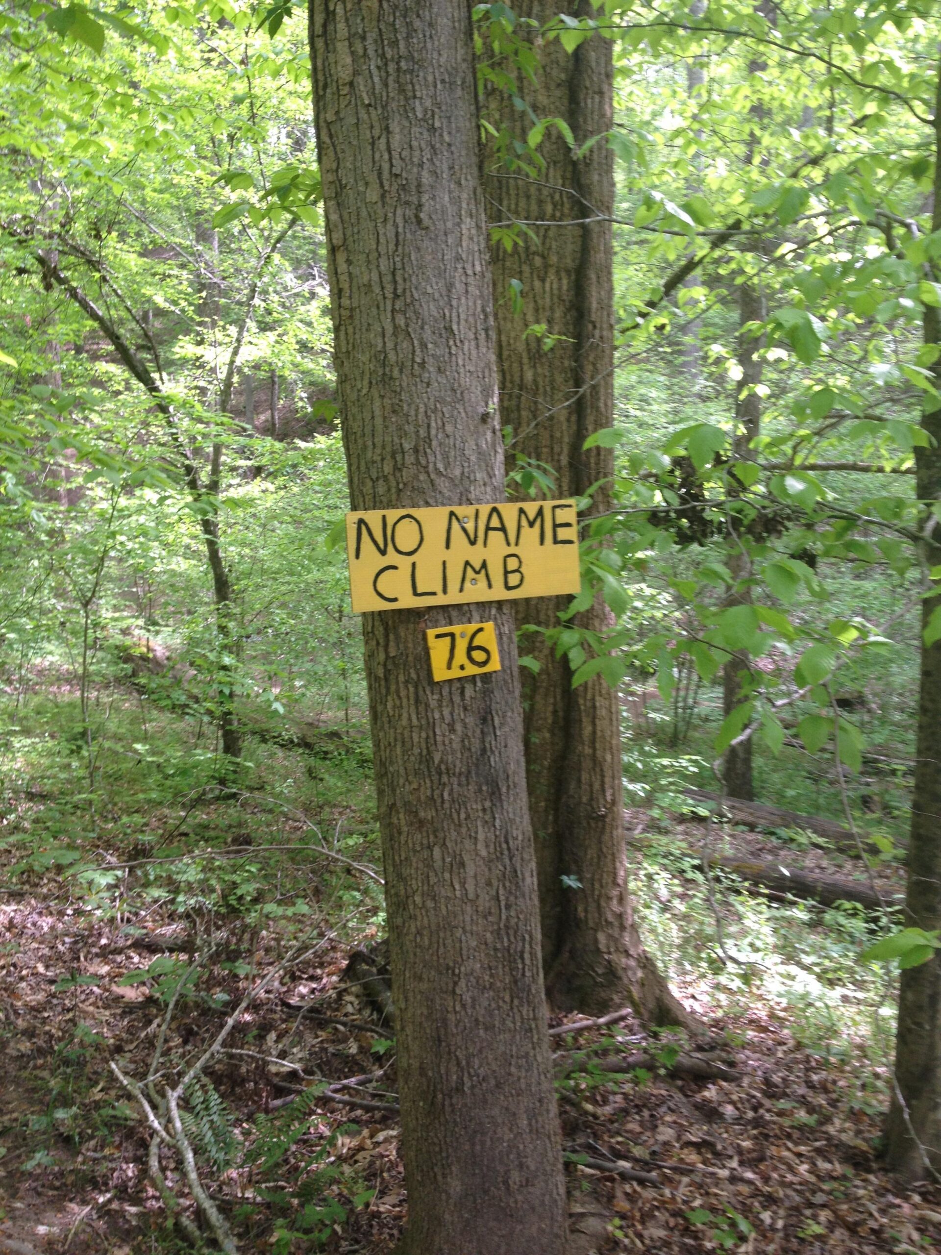 A yellow sign attached to a tree in a forested area reads "NO NAME CLIMB," with the number "7.6" displayed beneath it. The surrounding environment features lush green foliage and a natural landscape. Rocky River Trail mountain bike trail.
