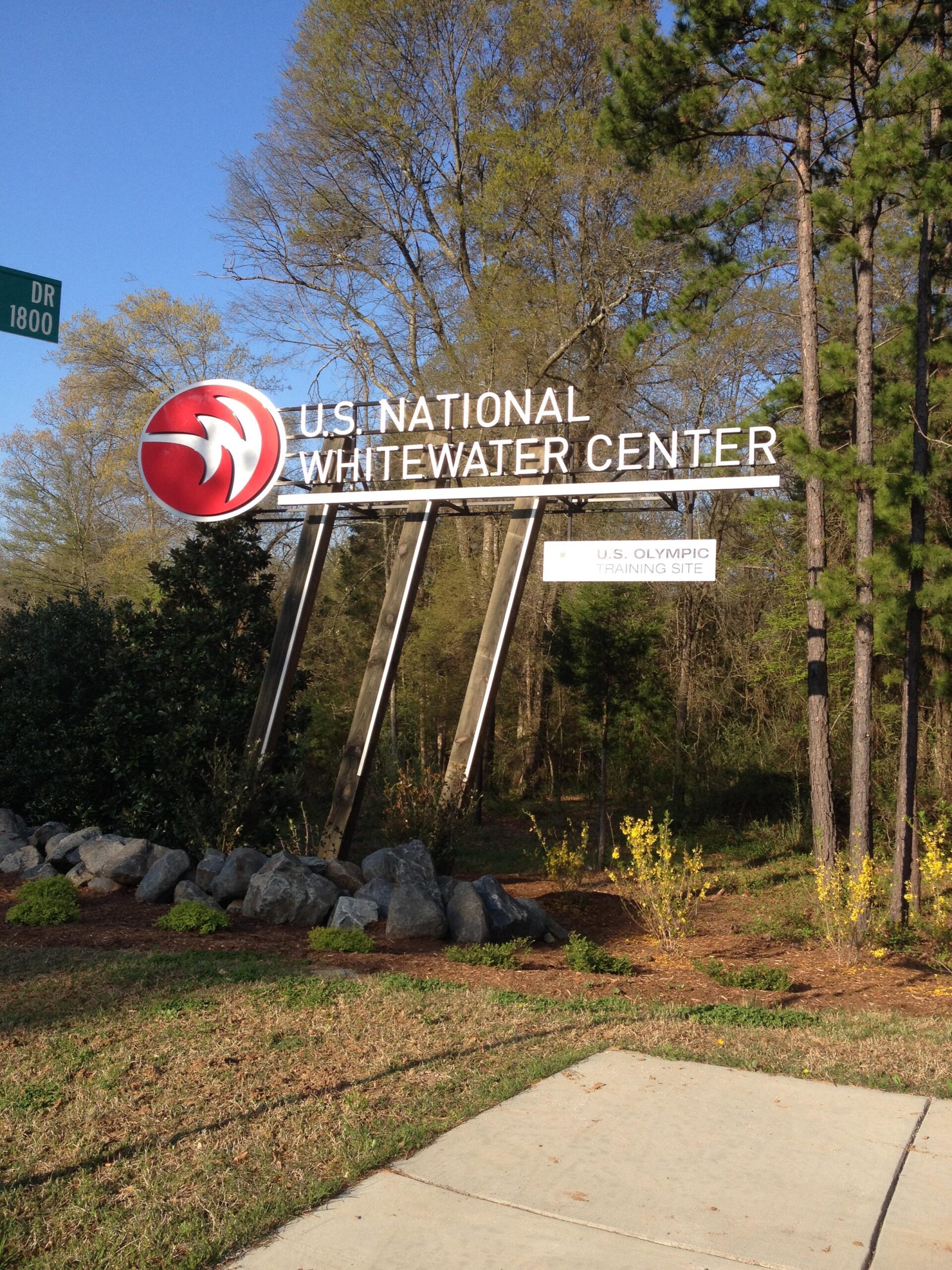 Sign for the U.S. National Whitewater Center, featuring a prominent logo and text indicating its status as a U.S. Olympic Training Site, surrounded by trees and landscaping. USNWC mountain bike trail.
