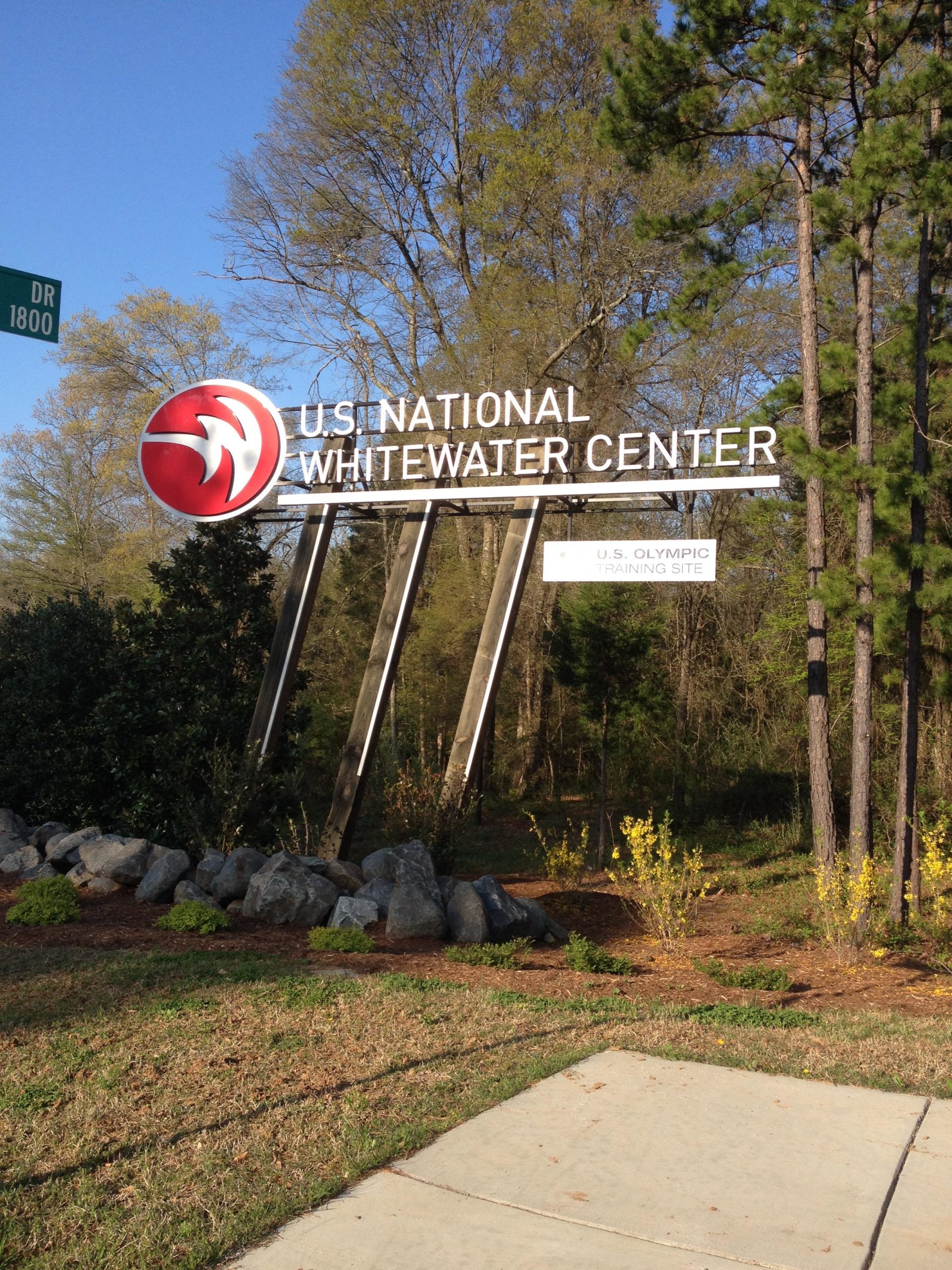 Sign for the U.S. National Whitewater Center, featuring a large logo and text indicating it is a U.S. Olympic training site. The background includes trees and landscaping. USNWC mountain bike trail.