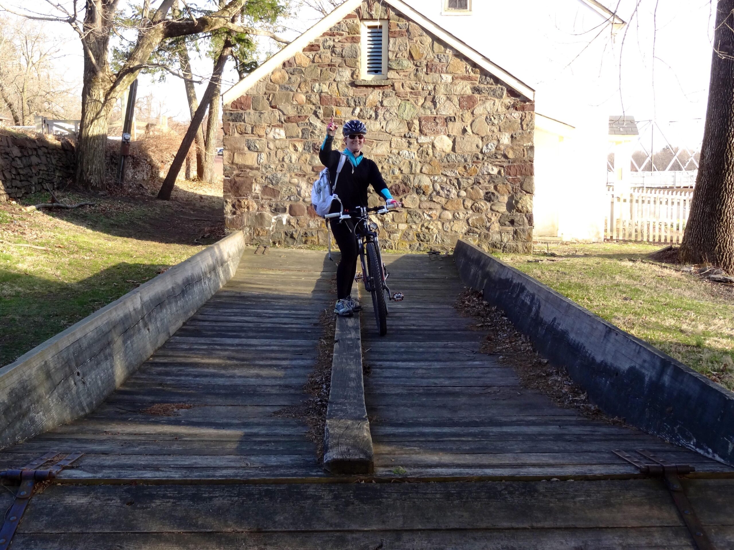 A person wearing a helmet and casual clothing is standing next to a bicycle on a wooden ramp leading up to a stone building. The individual is waving and appears to be enjoying the outdoor setting, with trees and grass in the background. The scene is illuminated by natural light, indicating a clear day. Pemberton Rail Trail mountain bike trail.