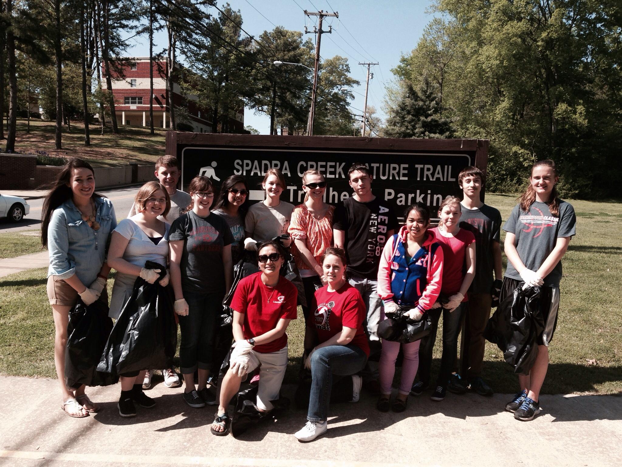 A group of diverse individuals posing in front of a sign that reads "Spadra Creek Nature Trail." They are wearing casual clothing, some holding large black garbage bags, suggesting they are participating in a cleanup activity. Lush trees and a grassy area are visible in the background, indicating a natural setting. Spadra Creek Nature Trail mountain bike trail.