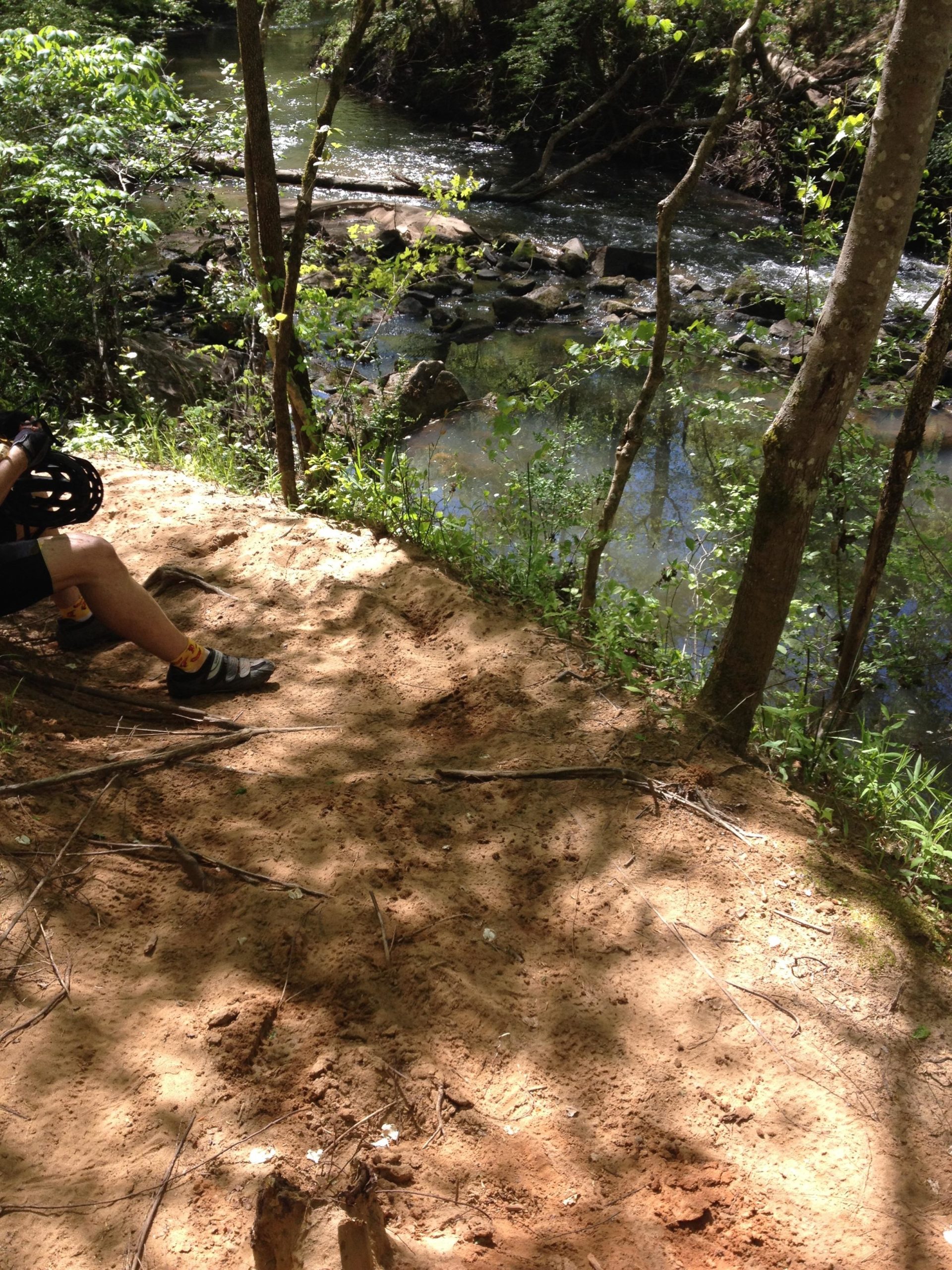 A sunlit view of a sandy riverbank surrounded by trees, with a gently flowing stream nearby. The image shows a portion of a person's leg and foot in black sandals, sitting on the edge of the bank. Small rocks and logs can be seen in the water, along with greenery along the shore. Anne Springs Close Greenway mountain bike trail.