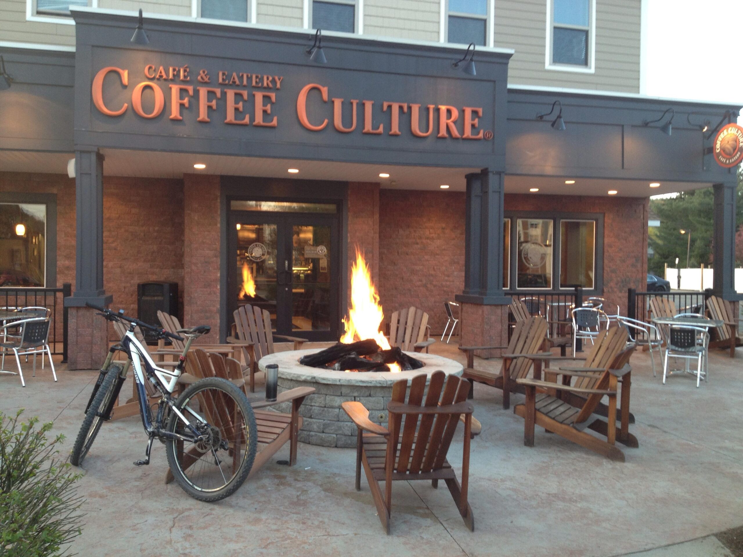 Specialized Stumpjumper FSR Comp 29: A cozy café and eatery named "Coffee Culture," featuring a welcoming outdoor seating area with wooden Adirondack chairs around a fire pit. A mountain bike is parked nearby, and tables are set up on the patio. The entrance is marked by double doors and a warm, inviting glow from inside the café. The building has a modern yet rustic design, with brick and wood accents.