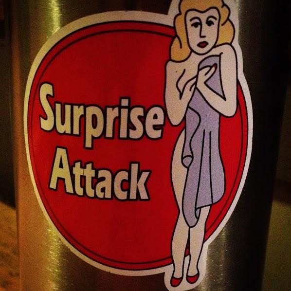 A round sticker featuring the text "Surprise Attack" in bold letters, accompanied by an illustration of a woman in a light purple dress with a vintage style, standing with one arm crossed over her chest and a playful expression. The background of the sticker is red, and it is applied to a metallic surface.