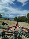 Cannondale Trail SL 29: Two red mountain bikes are positioned on a gravel path with a scenic view of a landscape in the background. The sky is partly cloudy, showcasing a mix of blue and white clouds, while trees are visible in the distance.