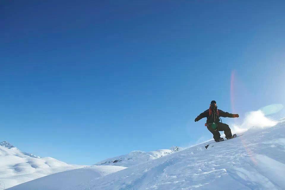 A snowboarder navigating a snowy slope against a clear blue sky, with mountains in the background. Snow is kicking up behind the boarder as they carve through the fresh powder.