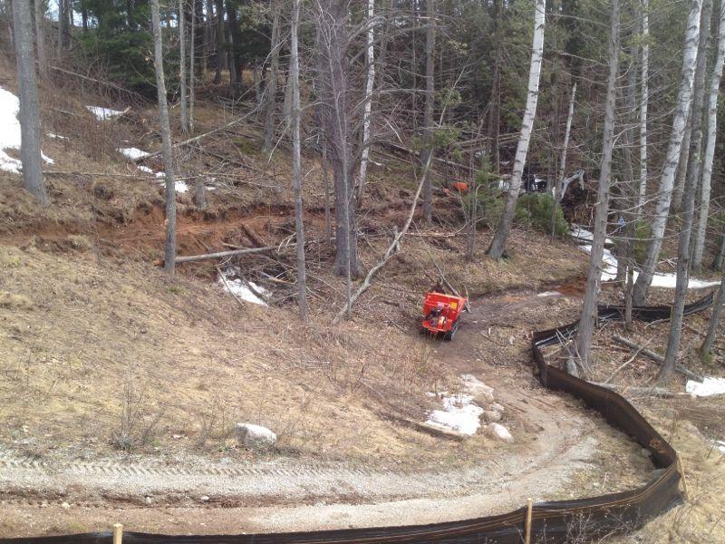 A construction site in a forested area, featuring cleared land with exposed soil and scattered logs. A small orange vehicle is visible near a winding dirt path, surrounded by trees with sparse foliage and remnants of snow on the ground. A silt fence is erected along the edge of the clearing. Noquemanon Trails Network: South Marquette Trails mountain bike trail.