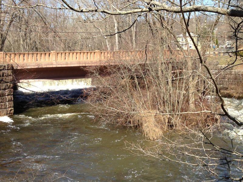 A wooden bridge spans over a flowing river, surrounded by bare trees and shrubs. The water below is slightly turbulent, indicating a recent rainfall or snowmelt. In the background, a glimpse of a building is visible through the trees, suggesting a rural setting. Wadsworth State Park mountain bike trail.