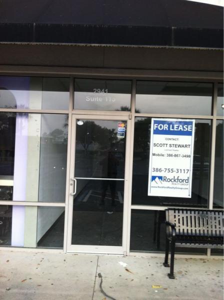 Front entrance of a commercial space for lease, featuring a sign with contact information for leasing inquiries. The glass door has a small "Open" sticker, and a bench is placed in front of the entrance. The suite number is displayed as 113, and the address is visible above the door.