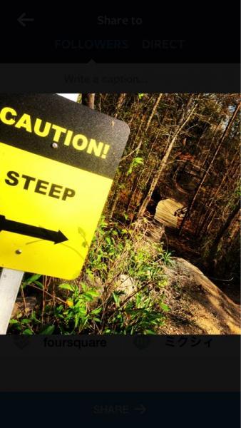 A bright yellow caution sign reads "CAUTION! STEEP" with an arrow pointing to the left. The sign is positioned on a narrow trail surrounded by trees and bushes, indicating a steep area ahead. In the background, a visible path leads down, hinting at the terrain's steepness. Horry County Bike Run Park mountain bike trail.