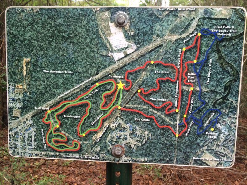 A map displaying various trails in a woodland area, including trail names like "Cambodia," "The Bone," and "Treehugger." The map features marked paths in red, blue, and green, with labeled parking and access points. The surrounding area includes trees and natural forest elements. UWF Mountain Bike Trails mountain bike trail.