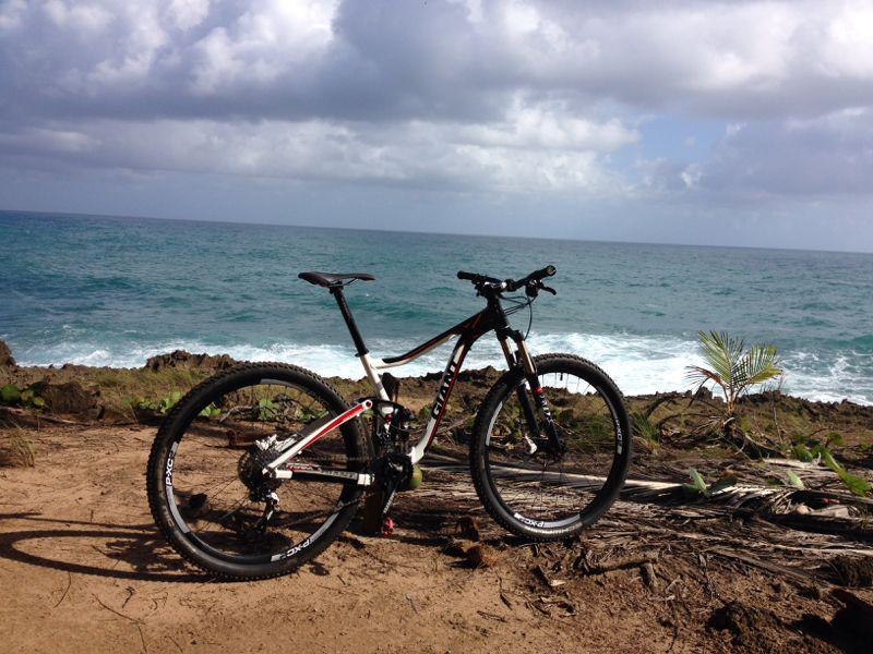 A mountain bike parked near a rocky shoreline, with the ocean visible in the background. The scene features blue water and partly cloudy skies, creating a scenic outdoor environment. Cerro Gordo Trail mountain bike trail.