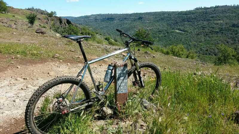 A mountain bike rests against a trail marker on a rocky hillside, surrounded by lush greenery and a scenic view of distant hills. The marker indicates trail directions and distances, with a clear blue sky overhead. "B" mountain bike trail.