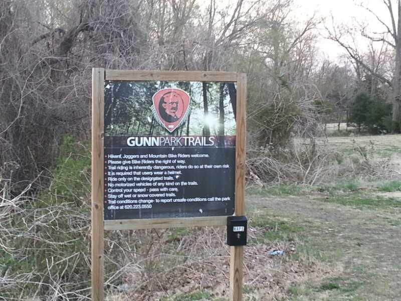 Sign for Gunn Park Trails, featuring safety and usage guidelines for hikers, joggers, and mountain bike riders. The background shows a natural setting with trees and open land. Gunn Park Trails mountain bike trail.