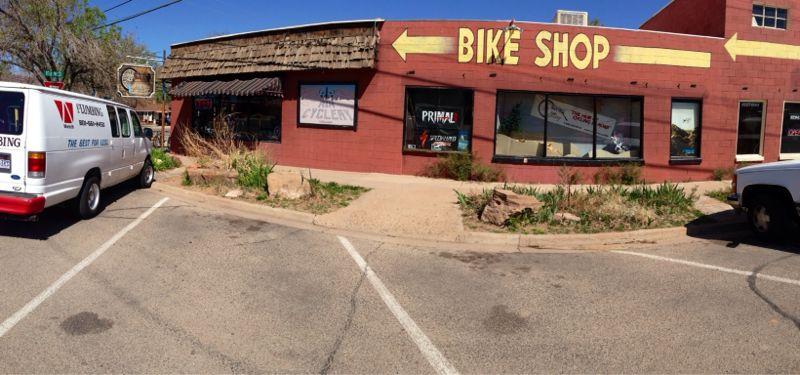 A panoramic view of a bike shop with a large yellow sign pointing left, indicating the direction of the entrance. The shop features windows displaying various biking accessories and apparel. A white van is parked in front of the shop, along with another vehicle, with a clear blue sky in the background. The landscape includes some greenery and unpaved areas adjacent to the building.
