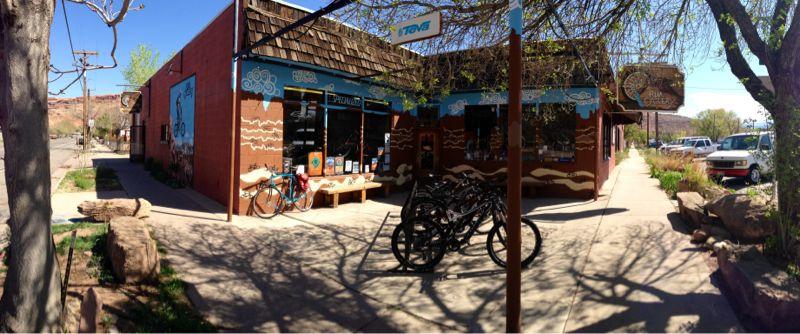 A vibrant street scene featuring a colorful storefront with outdoor seating and bicycles parked outside. The building is adorned with artistic murals, and trees provide shade along the sidewalk. In the background, cars are parked along the street, and the surrounding landscape shows hints of natural beauty.