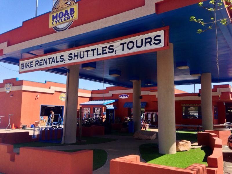 Exterior of Moab Cyclery featuring a bright blue and red building. A large sign reads "BIKE RENTALS, SHUTTLES, TOURS." The area is landscaped with grass and rocks, and bikes are visible in the background.