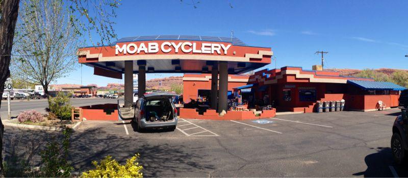 Exterior view of Moab Cyclery, a bike shop featuring a distinctive red and blue building. The image shows a parking area with a vehicle parked in front, surrounded by trees and shrubs. The backdrop includes rocky terrain typical of the Moab area under a clear blue sky.