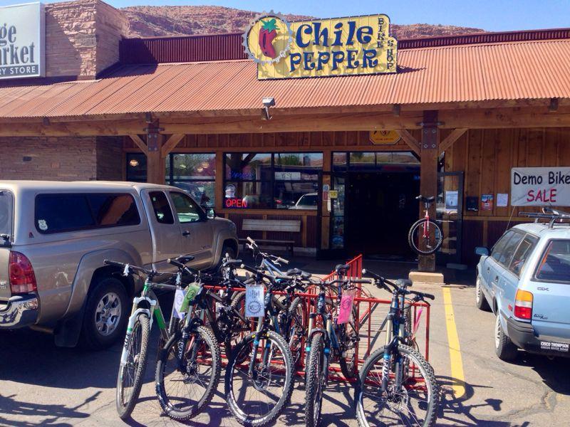 Exterior view of the Chile Pepper bike shop, featuring a wooden facade with a red metal roof. Several bicycles are parked in front of the entrance, which displays an "OPEN" sign and a sale notice for demo bikes. Two vehicles are parked nearby, set against a backdrop of mountains under a clear blue sky.