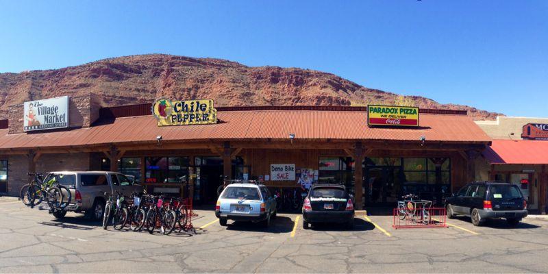 A sunny view of a commercial plaza featuring several storefronts, including "Chile Pepper" and "Paradox Pizza." The area is bustling with parked bicycles and vehicles, with a backdrop of a rocky mountain. Bright blue sky overhead and a sign advertising a "Demo Bike SALE" can also be seen.