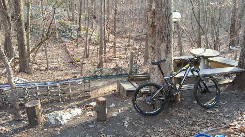 A mountain bike is parked next to a wooden table and chairs in a wooded area, surrounded by bare trees and rocky terrain. A small dirt path winds through the scene, leading away from the bike towards a bridge-like structure in the background. The ground is covered with fallen leaves, indicating an early spring or late autumn setting. Needham Town Forest mountain bike trail.