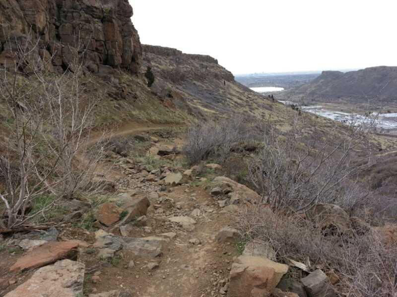 A rocky hiking trail winding through a hilly landscape, with sparse vegetation and a view of a river and distant hills in the background under a cloudy sky. North Table Mountain mountain bike trail.