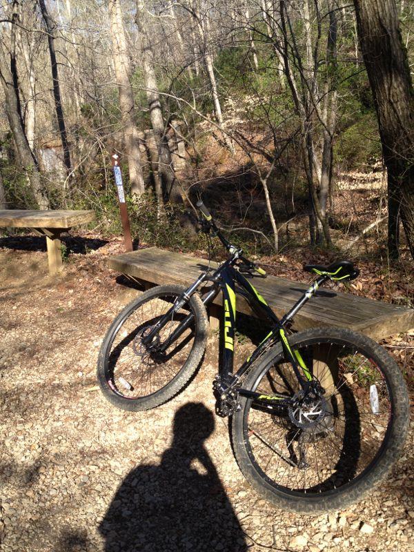 A mountain bike leaning against a wooden bench on a gravel trail surrounded by trees, with a trail sign visible in the background. Sunlight filters through the branches, creating a natural outdoor setting. Chicopee Woods mountain bike trail.