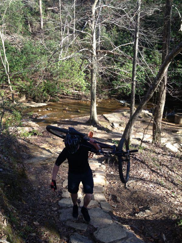 A person wearing a helmet and cycling gear carries a mountain bike up a rocky trail beside a creek in a wooded area. The scene features bare trees and green foliage, indicating an outdoor adventure. Chicopee Woods mountain bike trail.