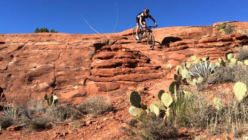 A mountain biker navigating a rocky terrain with red sandstone and cacti in the foreground under a clear blue sky. Hiline mountain bike trail.